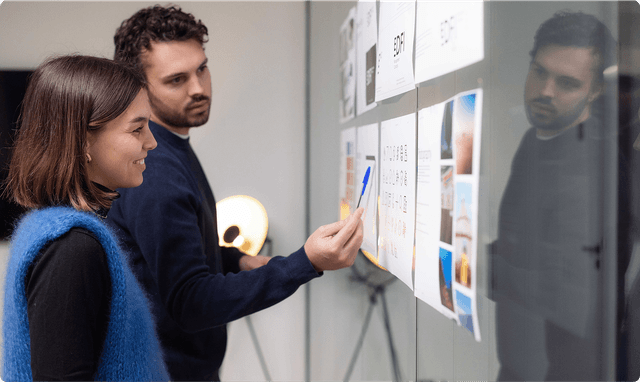 Brand Identity Monitoring Two people discussing and pointing at documents on a glass wall, with a lamp in the background.
