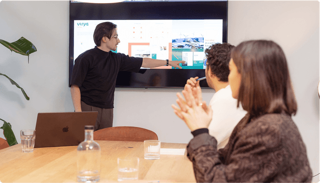 Brand identity design A person presents slides on a screen to two seated individuals in a meeting room with a wooden table and a plant in the corner.