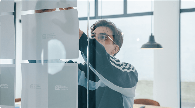 Stylescape Person in glasses and a striped sweater attaching papers to a glass wall in a bright room with modern decor.