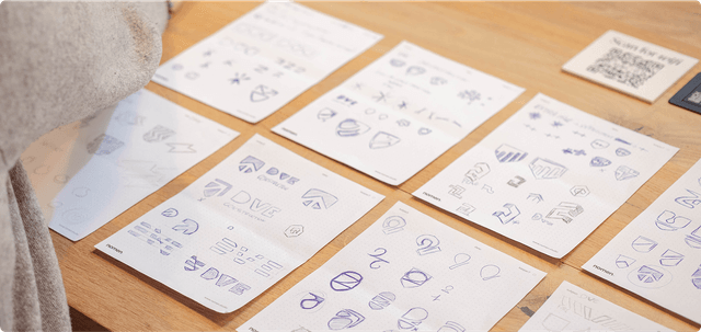 Brand book Sheets of paper on a wooden table displaying various hand-drawn logo sketches and designs in blue ink.