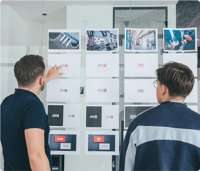 Brand identity design Two people examining a wall display of printed images and designs, discussing various layouts in a modern office setting.