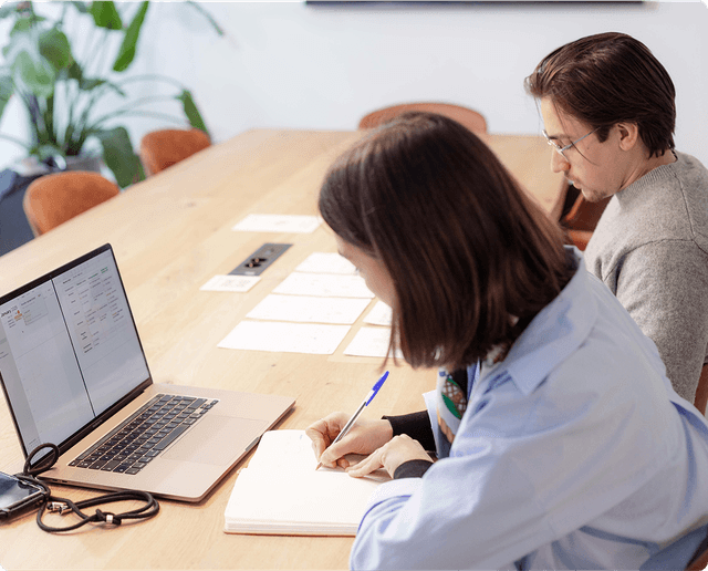 Brand Identity Monitoring Two people working at a wooden table with a laptop, papers, and notebooks. One is writing, the other is focused on the screen.