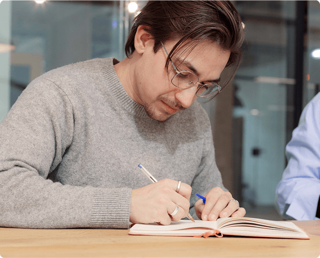 Brand Identity Monitoring A person with glasses and a gray sweater writes in a notebook at a wooden table in a modern office setting.