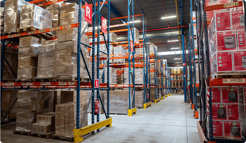 Warehouse interior with tall shelves filled with stacked boxes and pallets. A clear aisle runs down the center, under bright overhead lighting.