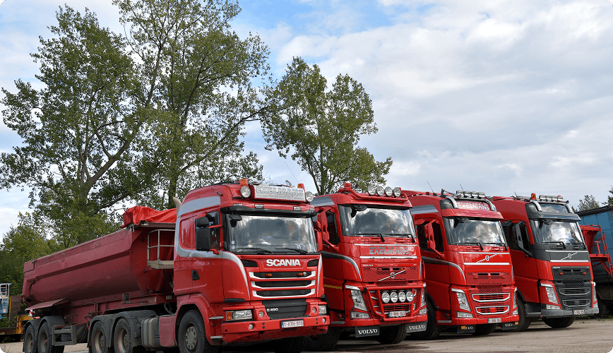 Five red semi-trucks parked in a row on a clear day, with trees and a cloudy sky in the background.
